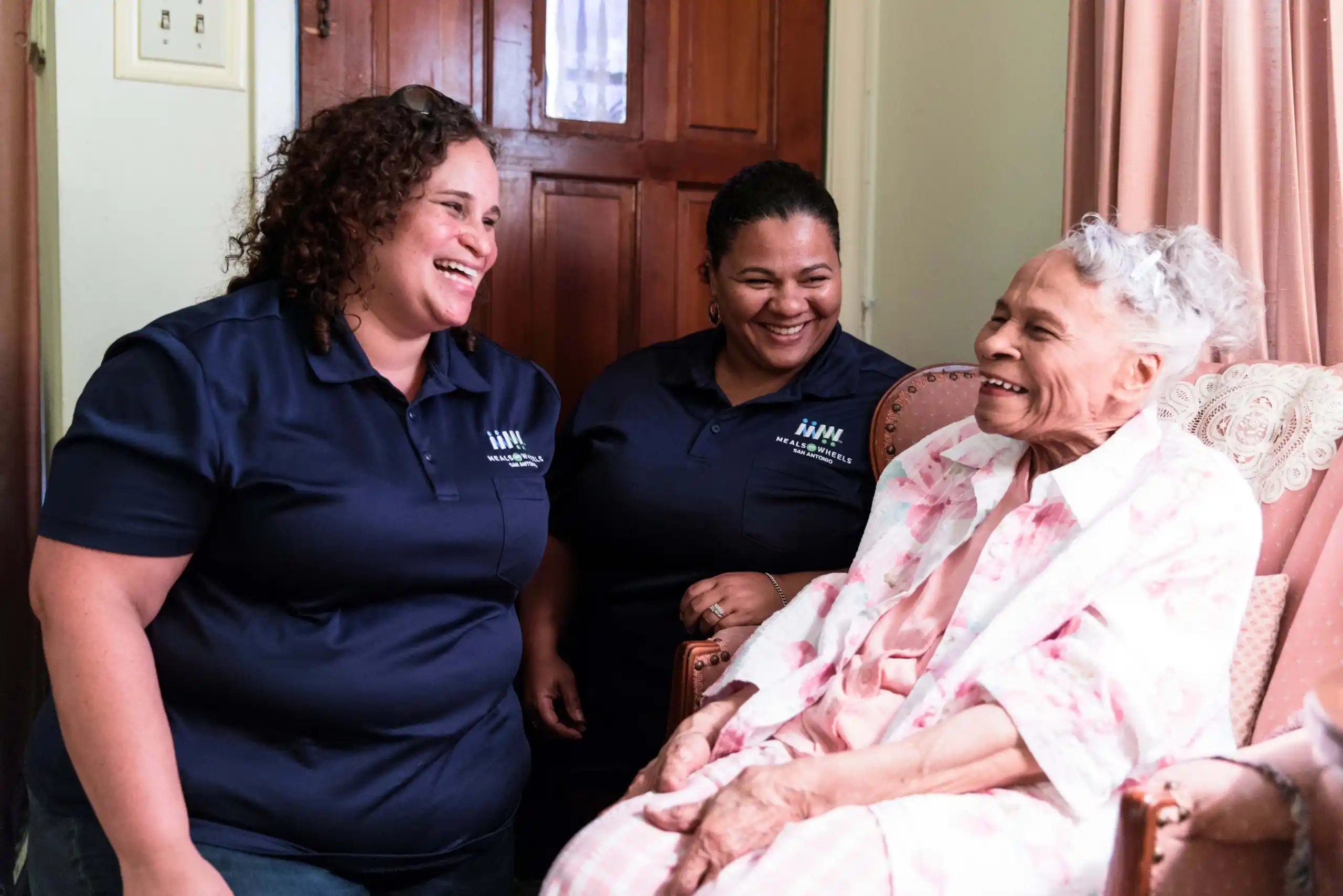 Two women volunteers standing by a smiling senior sitting in a chair.
