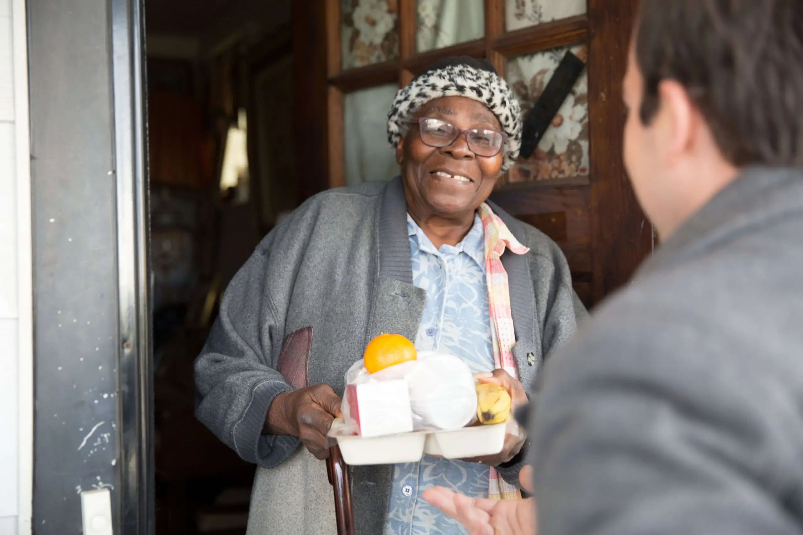 Black female senior smiling and talking to volunteer while holding a meal at front door.