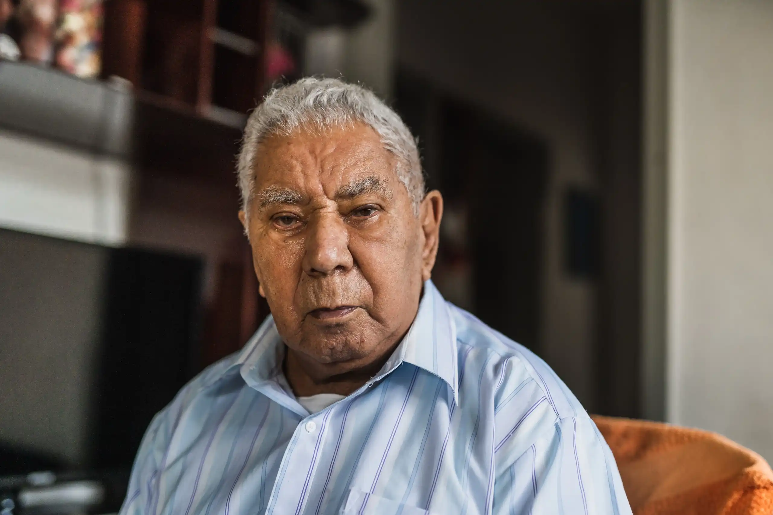 Senior male sitting alone looking somber while wearing striped blue shirt.
