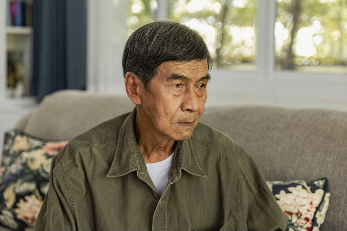 Asian male senior sitting alone on couch looking off into the distance.