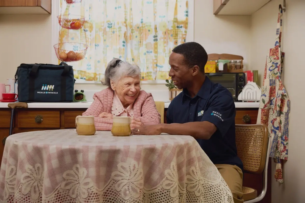 White female senior and Black male volunteer sitting together at a kitchen table smiling.