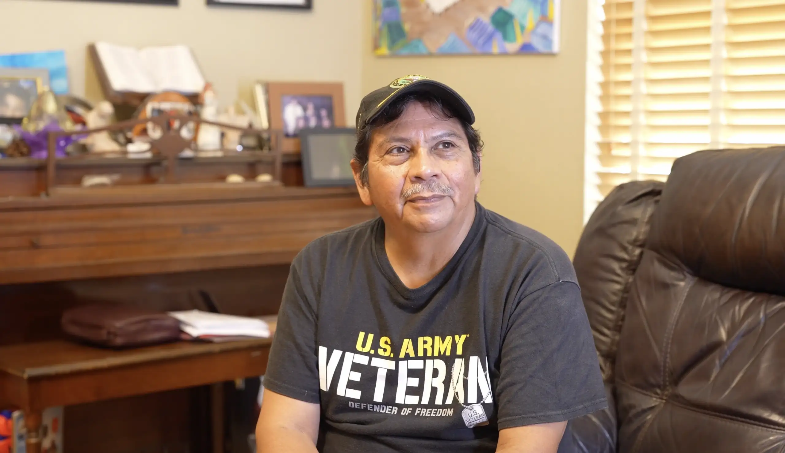 Veteran Juan Lopez sitting on couch wearing a U.S. Army Veteran shirt and a hat.