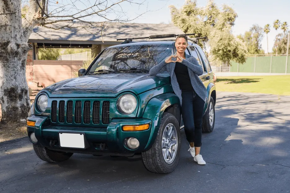 Black female standing in front of a teal car holding her hands in the shape of a heart.