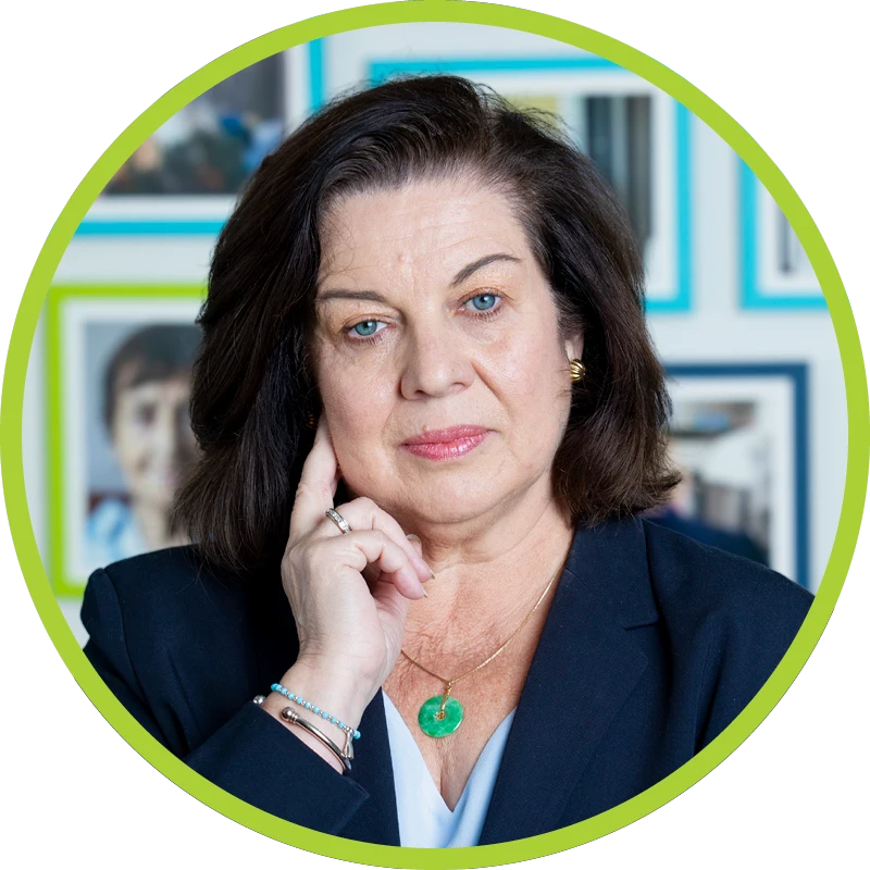 Deirdre McGinley Geiser smiling wearing a light blue top and navy-blue blazer, gold earrings, green jade necklace, bracelets, and a ring.