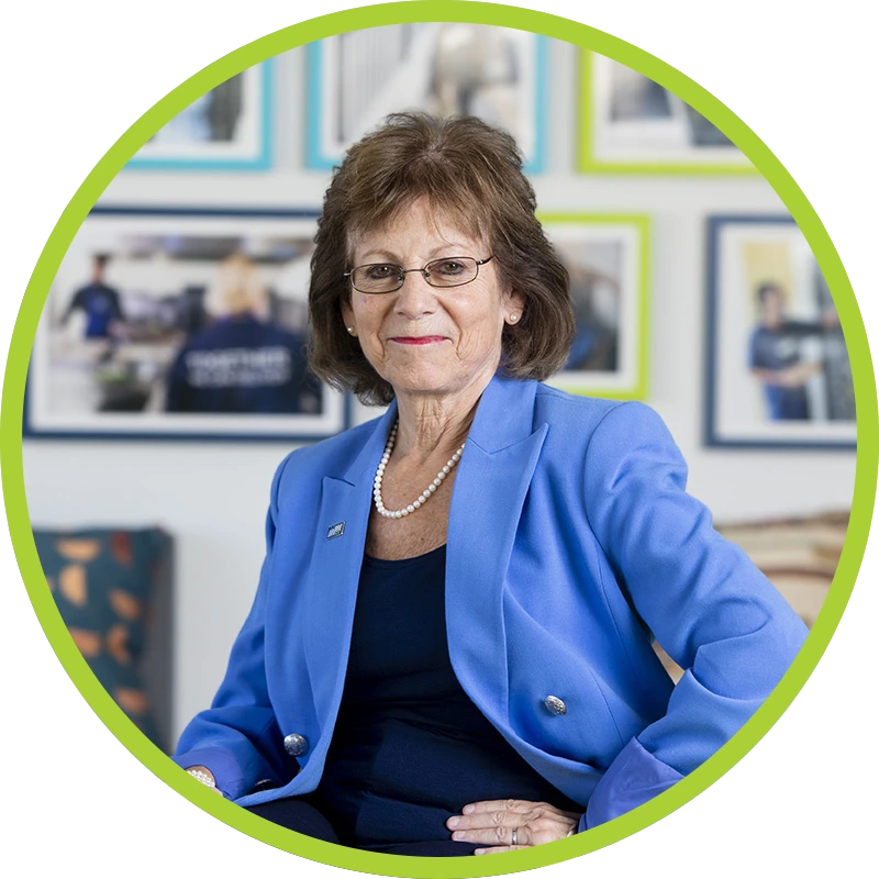 Ellie Hollander smiling wearing glasses, pearl necklace, light blue blazer and navy top and bottoms.
