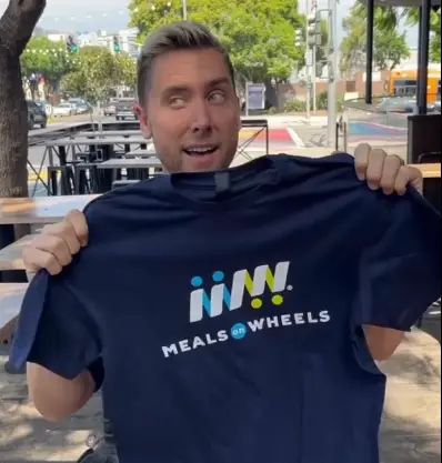 Lance Bass holding up a navy-blue Meals on Wheel shirt.