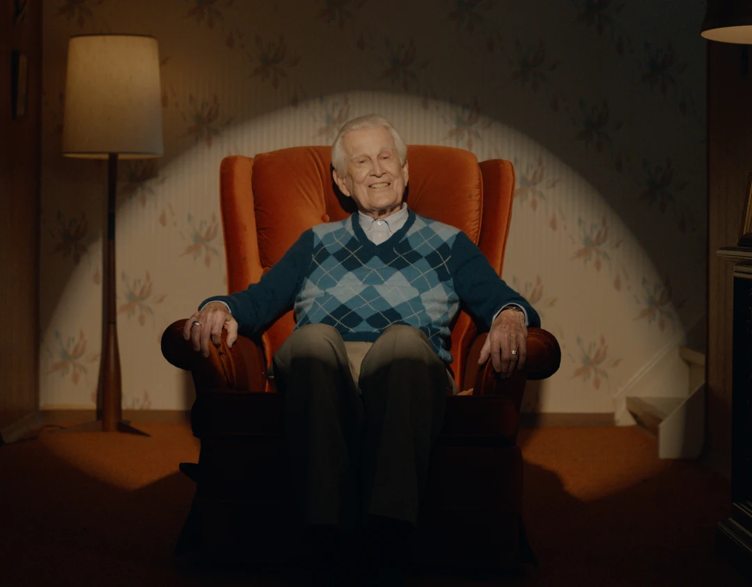 White male senior Lloyd sitting on an armchair smiling with a spotlight surrounding him.