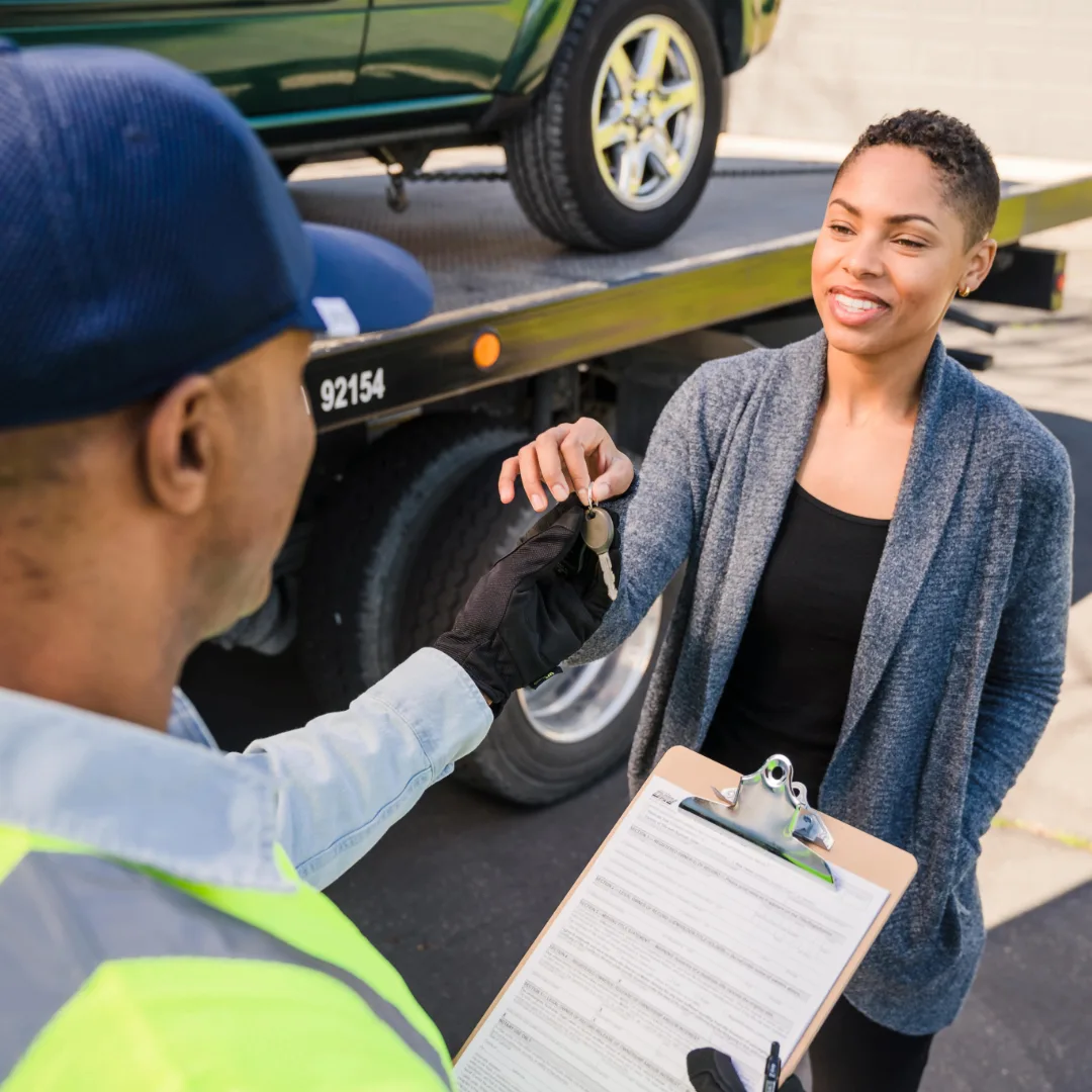 Black male handing car key to Black female while standing next to a car on a tow truck.