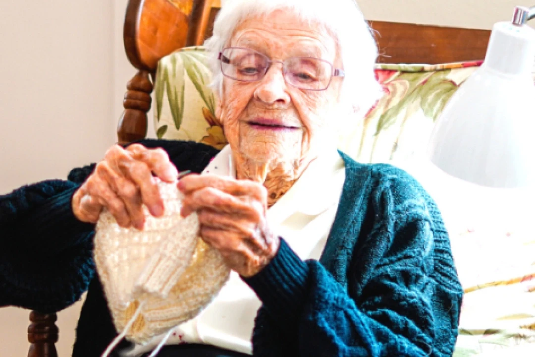Client, Gundy, smiling and knitting on bed.