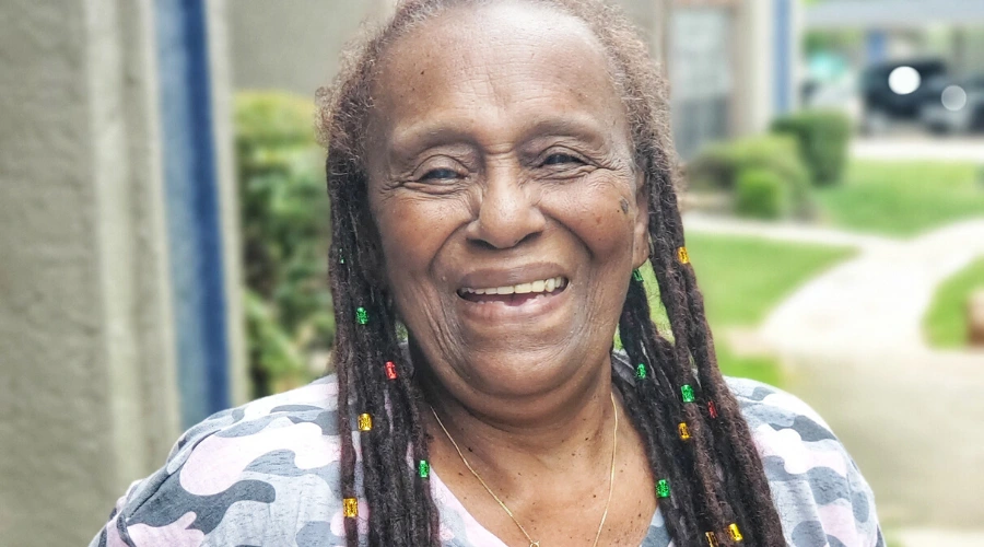 Shirley, a Meals on Wheels client in Dallas, TX smiling while wearing a pink camouflage top and locs in her hair.