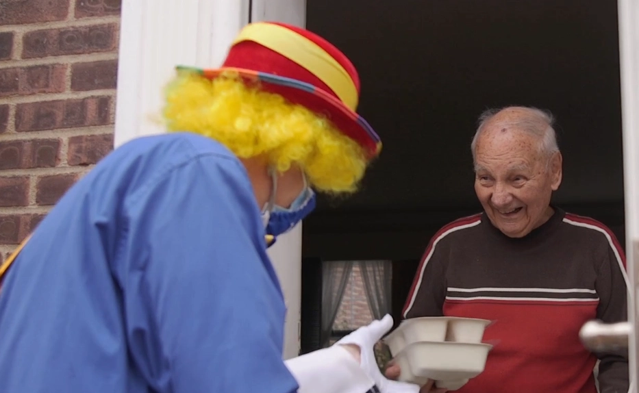 Macy's volunteer dressed as a clown delivering meals to a senior male.