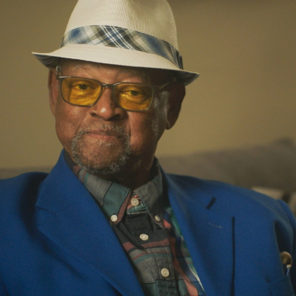 Meals on Wheels Client Client, Wilbert, Wearing Blue Blazer Maintaining Eye Contact and Sitting Smiling on Couch.