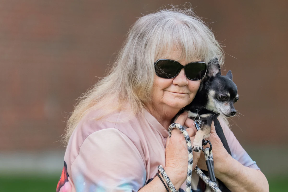 Older adult female smiling while squatting in grass holding her Chihuahua-pinscher mix dog Max.